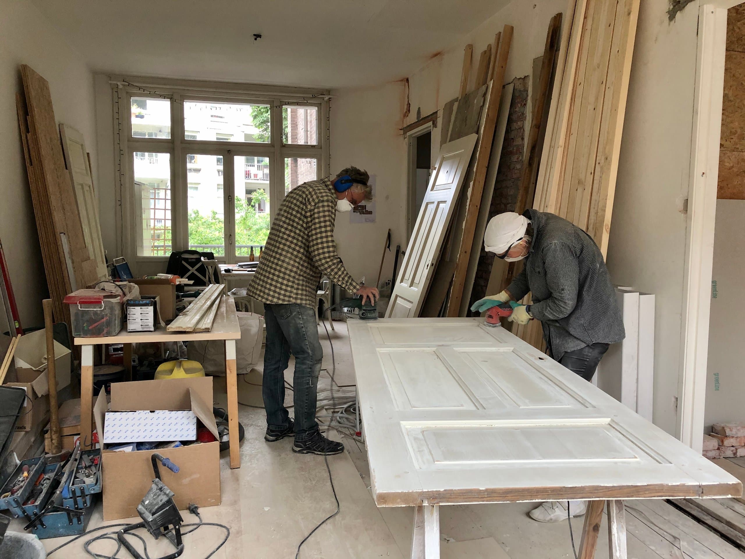 Twee mensen schuren een paneeldeur op een werktafel midden in de verbouwing