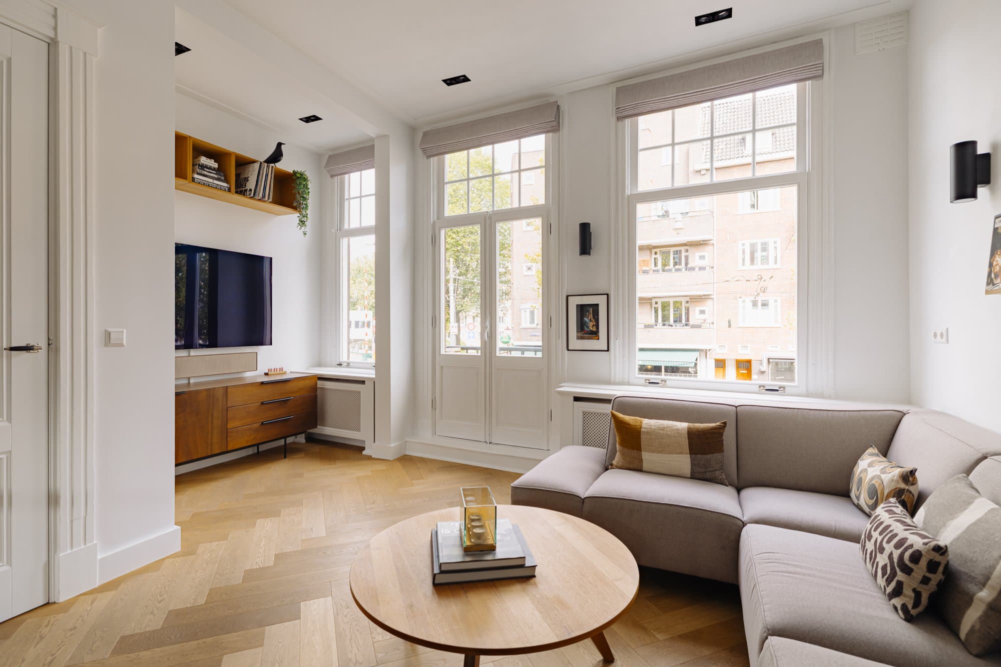 De woonkamer met houten tv-meubel onder de hoge ramen